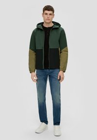 Kleurblokjas in groen en khaki, met een capuchon, ritssluiting en elastische manchetten. Combinatie met blauwe jeans en witte sneakers.