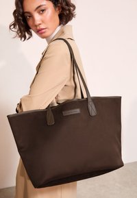 Bruine draagtas gemaakt van zacht materiaal, met stevige handvatten, een label met "friends like them", en zonder zichtbare patronen of accenten.