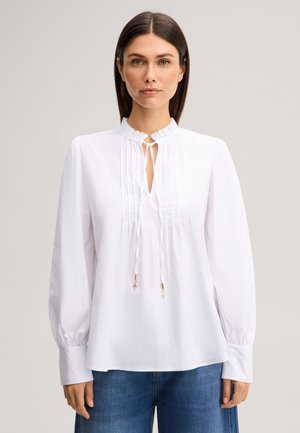 Femme aux cheveux bruns et raides porte un chemisier blanc à manches longues avec des plis et des lacets, associé à un jean bleu, sur un fond uni.
