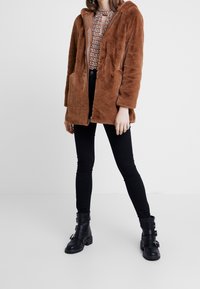 Brown faux fur jacket with a hood, front pockets, and zip closure, worn over a patterned top and paired with black skinny jeans and black boots.