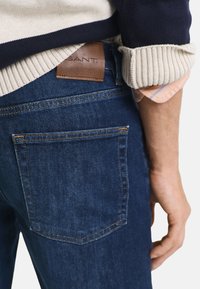 Jeans in denim blu scuro con una texture liscia. Presenta un'etichetta in pelle marrone con la scritta "SANT" e un design a cinque tasche. Polsini a coste della maglia visibili.