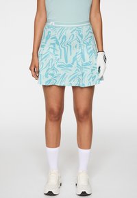 Donna che indossa una gonna plissettata blu con motivi, scarpe sportive bianche, calzini bianchi e un guanto da golf bianco sulla mano destra.