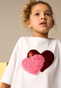 White short-sleeved t-shirt featuring two heart designs: a shiny red sequin heart and a fluffy pink heart. Relaxed fit.