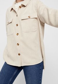 Beige kordfluweel skjorta med knappar och två framfickor, bärs över en vit topp och matchas med mörkblå jeans.