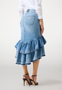 Vrouw in een lichtblauwe, gerimpelde denim midi-rok, een witte blouse met lange mouwen en zwarte hoge hakken, staande tegen een lichte achtergrond.