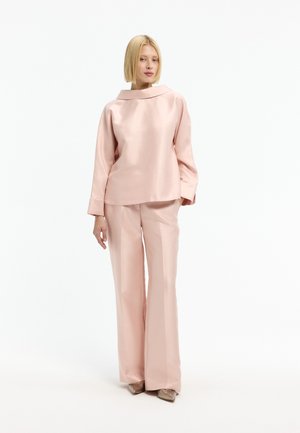 Frau trägt eine locker sitzende, blassrosa Satinbluse mit weitem Kragen und passende weit geschnittene Hosen, steht vor einem weißen Hintergrund.