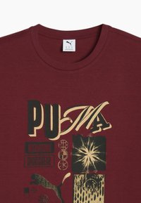Maroon Puma t-shirt with graphic text "PUMA," "International Sportswear," abstract shapes, and puma logo in black and beige tones.