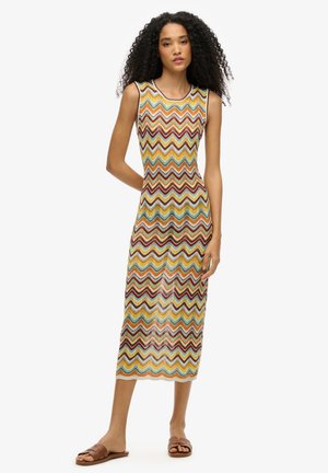 Gebreide mouwloze jurk met een zigzagpatroon in rood, geel en groen, met een slim fit en een lengte tot halverwege de kuit. Bruine platte sandalen worden gedragen.