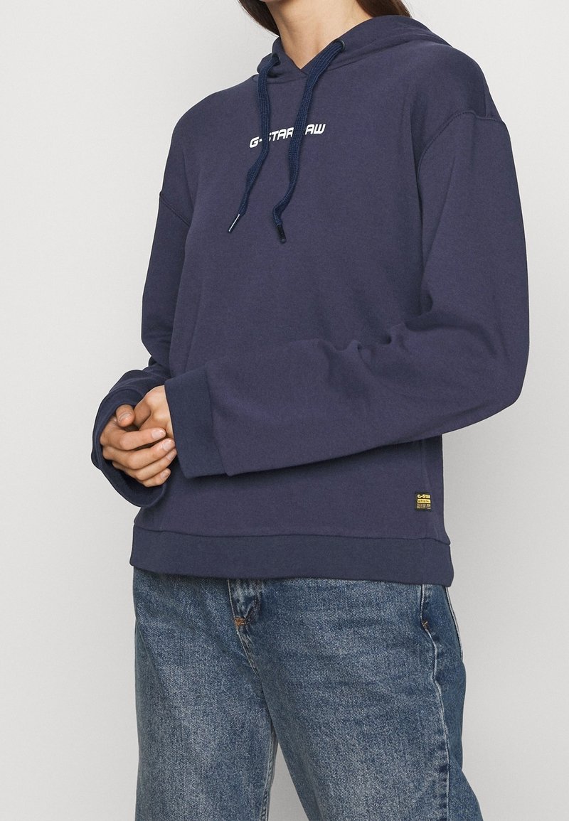 Personne portant un sweat à capuche bleu marine G-Star Raw avec cordons et un jean bleu délavé, mains jointes devant.