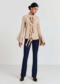 Blusa beige con frontale a rouches e maniche a campana, abbinata a jeans svasati blu scuro. Il tessuto appare liscio e leggero.