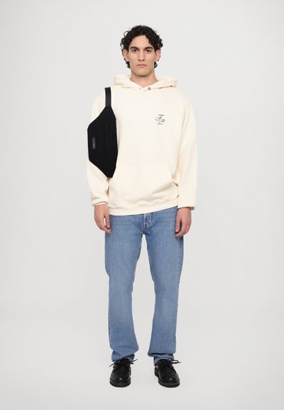 Light beige hoodie with green logo, blue jeans, and black shoes. A black crossbody bag with a front zipper rests on the left shoulder.