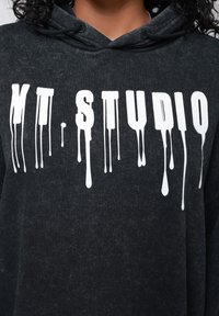 Čierna mikina s kapucňou s textúrovaným povrchom, na prednej strane s bielym nápisom „MT. STUDIO“ v štýle kvapkajúcej farby.