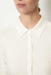 Femme portant une chemise à col crème à boutons, montrant le cou, les lèvres et le haut du torse sur fond clair.