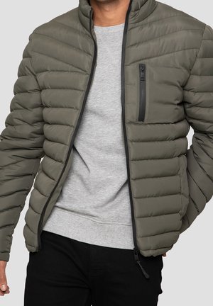 Homme portant une veste matelassée vert olive sur un sweat-shirt gris clair avec un pantalon noir, devant un fond uni.