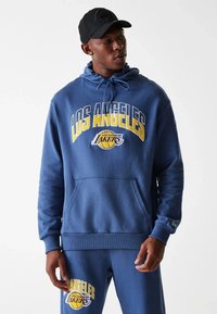 Sweat-shirt bleu avec "LOS ANGELES LAKERS" imprimé en jaune, doté d'une poche avant et de poignets côtelés. Associé à un pantalon bleu assorti.