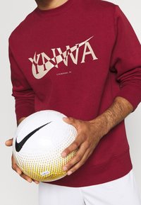 Personne portant un sweat-shirt bordeaux du Liverpool FC tenant un ballon de football Nike blanc et jaune avec un logo Swoosh noir.