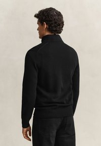 Hombre con cabello rizado y oscuro que lleva un suéter de cuello alto negro con textura acanalada y vaqueros negros, de pie de espaldas frente a un fondo liso.