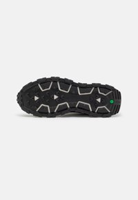 Semelle de chaussure noire robuste avec des rainures profondes, des accents blancs angulaires et un petit hexagone vert près du talon sur fond blanc.