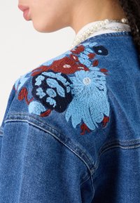 Jacket dżinsowy z intensywnym niebieskim i czerwonym haftem w kwiaty na ramieniu. Klasyczny krój z teksturowanym materiałem i starannym szyciem.