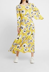 Abito lungo giallo a fiori con maniche lunghe, caratterizzato da un motivo di fiori bianchi e neri, con scollatura arricciata e silhouette fluida.