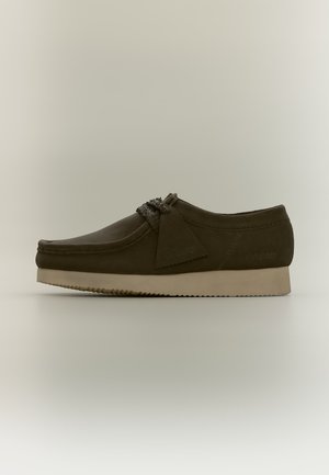 Brauner Wildleder-Mokassinschuh mit cremefarbener Sohle und dekorativer Quaste auf neutralem Hintergrund, Seitenansicht.