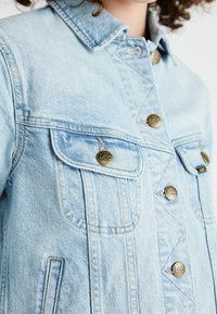 Ανοιχτό γαλάζιο τζάκετ από τζην με κλασικό γιακά, που διαθέτει χάλκινους κουμπιά και δύο μπροστινές τσέπες. Το ύφασμα έχει μια διακριτική υφή.