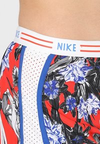 Sportovní kraťasy Nike s červeným květinovým vzorem a modrými akcenty. Pas je bílý s modrým nápisem a červenými pruhy. Boční panely jsou z texturované síťoviny.