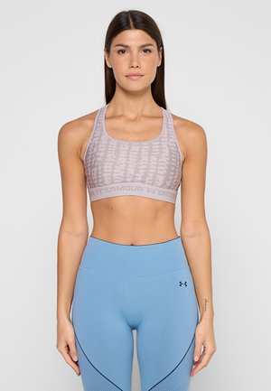 Femme portant un soutien-gorge de sport mauve clair à motifs et un legging bleu taille haute, debout devant un fond gris uni, regardant vers l’avant.