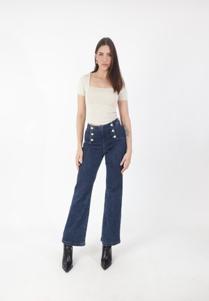Mujer de pie con el cabello castaño liso, vistiendo una blusa beige de escote cuadrado, jeans azules de tiro alto con botones y botas negras de punta.