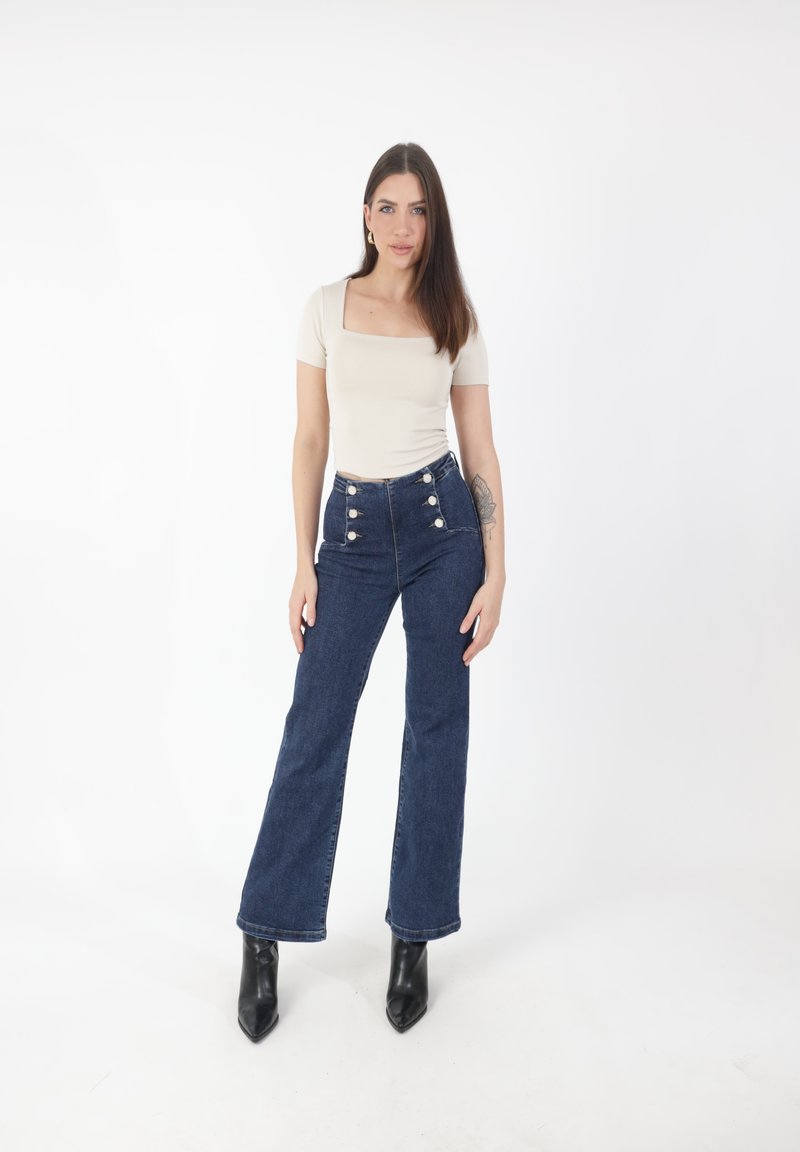 Donna in piedi con capelli castani lisci, indossa una maglietta beige a scollatura quadrata, jeans blu a vita alta con bottoni e stivali neri a punta.