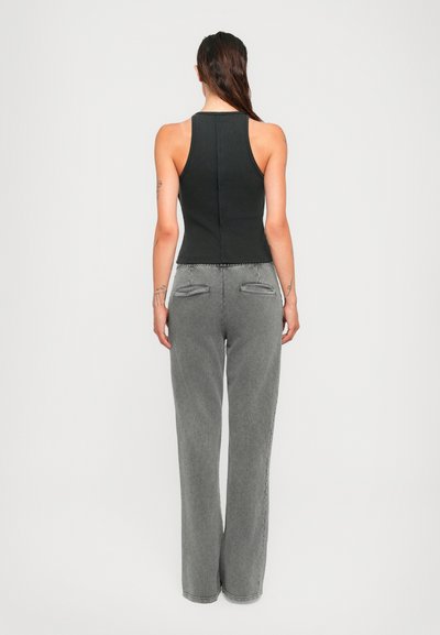 Woman with long dark hair wearing a sleeveless black top and high-waisted gray wide-leg pants, standing against a plain background.