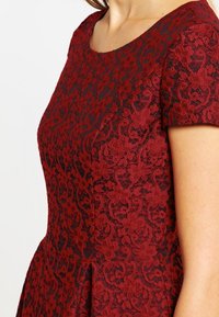 Femme portant une robe ajustée en dentelle rouge foncé avec des motifs floraux et des manches courtes, sur un fond blanc uni.