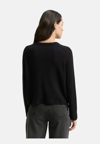 Frau mit langen braunen Haaren, die einen schwarzen Langarmpullover und dunkelgraue Jeans trägt, mit dem Rücken zu einem schlichten Hintergrund.