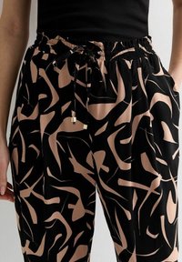 Vrouw in zwarte en beige abstracte patroonbroeken met elastische tailleband en gestrikt trekkoord, gecombineerd met een zwart mouwloos top.