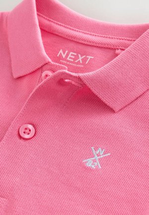 Roze poloshirt met een gestructureerde, geweven stof, twee bijpassende knopen en een wit geborduurd logo. Label met de tekst "NEXT, gemaakt in Bangladesh."