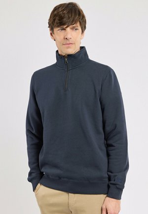 Homme portant un pull bleu marine à fermeture éclair au quart et un pantalon beige, debout les mains dans les poches, regardant légèrement sur le côté.
