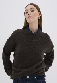 Brauner Strickpullover mit hohem Kragen, lockerer Passform und weicher Textur, über einem Denimhemd getragen und kombiniert mit dunkelblauen Jeans.