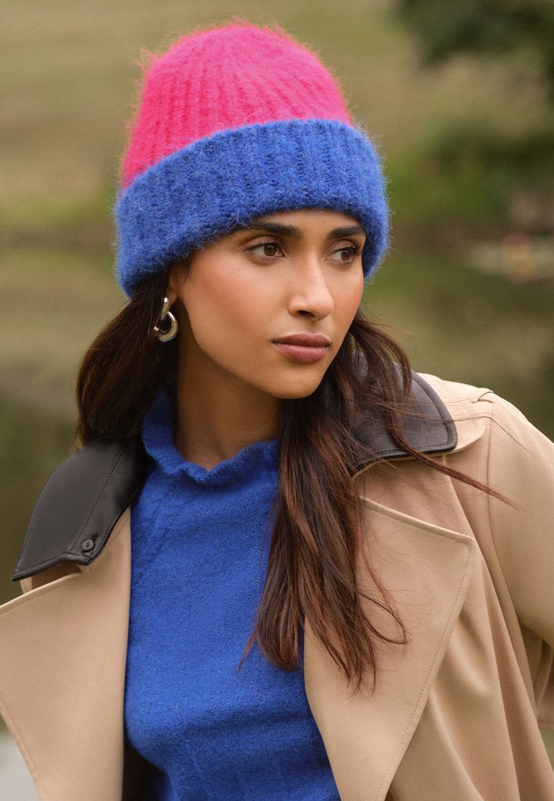 Strikket hue med lyserød top og blå bund; blød tekstur. Modellen har en blå top og en tanjakke med sort krave på. Sølvcreoler.