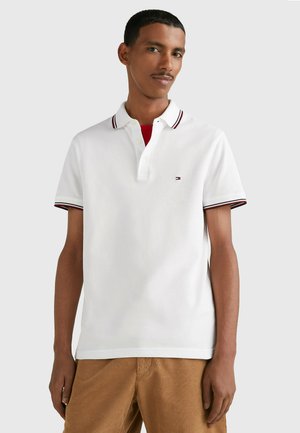 Polo bianco con pattina a tre bottoni, colletto a coste con strisce rosse e blu scuro, e un piccolo logo sul petto. Maniche corte.