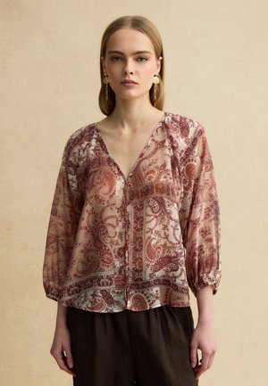 Frau trägt eine durchsichtige, langärmlige Bluse mit roten Paisley-Mustern und goldenen, hängenden Ohrringen, steht vor einem beigen Hintergrund.