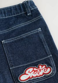 Tummansinisiä farkkuja, joissa on valkoiset tikkaukset ja suuri punainen brodeerattu "Skate" -patch takataskussa.