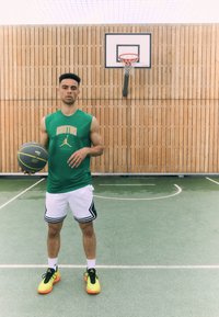 Green sleeveless basketball jersey with "BOSTON" text, white shorts with black stripes, and bright yellow-orange sneakers. Indoor court setting.