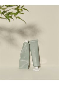 Kaksi pehmeää vihreää voidetuubia on asetettu pystysuoraan ja vaakasuoraan, ja niissä on minimalistista valkoista tekstiä sekä pieni kreemilattia neutraalilla pinnalla.