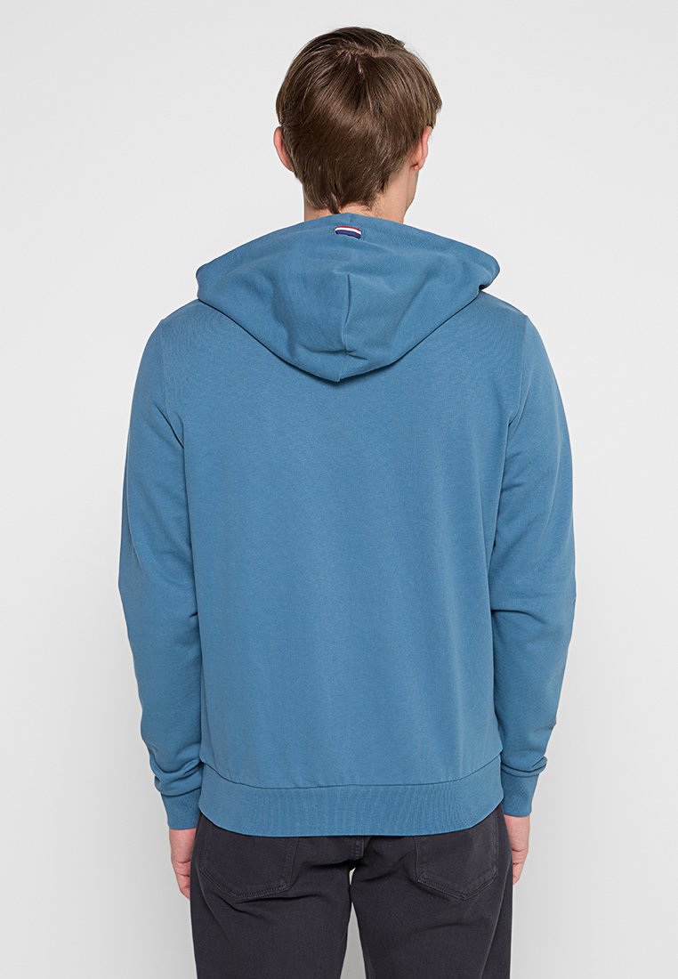 Mężczyzna w niebieskiej bluzie z kapturem stoi tyłem do kamery na jednolitym białym tle.