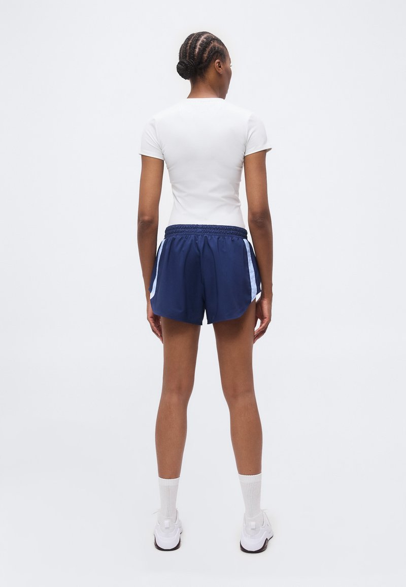 Mujer con cabello trenzado de espaldas, lleva una camiseta blanca de manga corta, pantalones cortos deportivos azul marino con rayas blancas, calcetines blancos y zapatillas blancas.