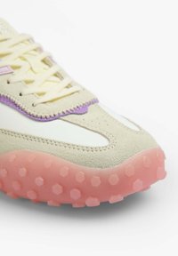 Beige sneaker with a textured suede upper, purple accents, and a pink rubber sole featuring hexagonal patterns for grip. Lace closure.