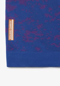 Blå stickad tyg med ett texturerat mönster i mörkare blått och rosa accenter; logotag i orange läser "Roland Garros" vid kanten.