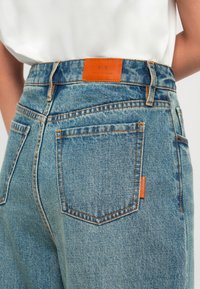 I jeans in denim blu presentano una vita alta, cuciture arancioni e un'etichetta in pelle arancione con "A/X" sul retro. Sono visibili due tasche posteriori.
