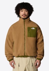 Giacca in fleece color marrone chiaro con accenti verdi, colletto alto, zip frontale e tasche laterali. Texture morbida; logo sulla tasca del petto.