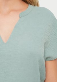 Top cu mâneci scurte, cu decolteu în V, realizat dintr-un material moale, texturat, de culoare verde deschis. Se remarcă printr-o croială lejeră, cu o linie de guler netedă și un model subtil de valuri.
